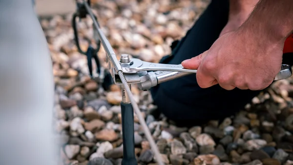 Nahaufnahme: Monteur befestigt EASiTec Seilsicherungssysteme auf Dächern mit Schraubenschlüssel an Edelstahlseil über Kiesfläche zur Absturzsicherung.