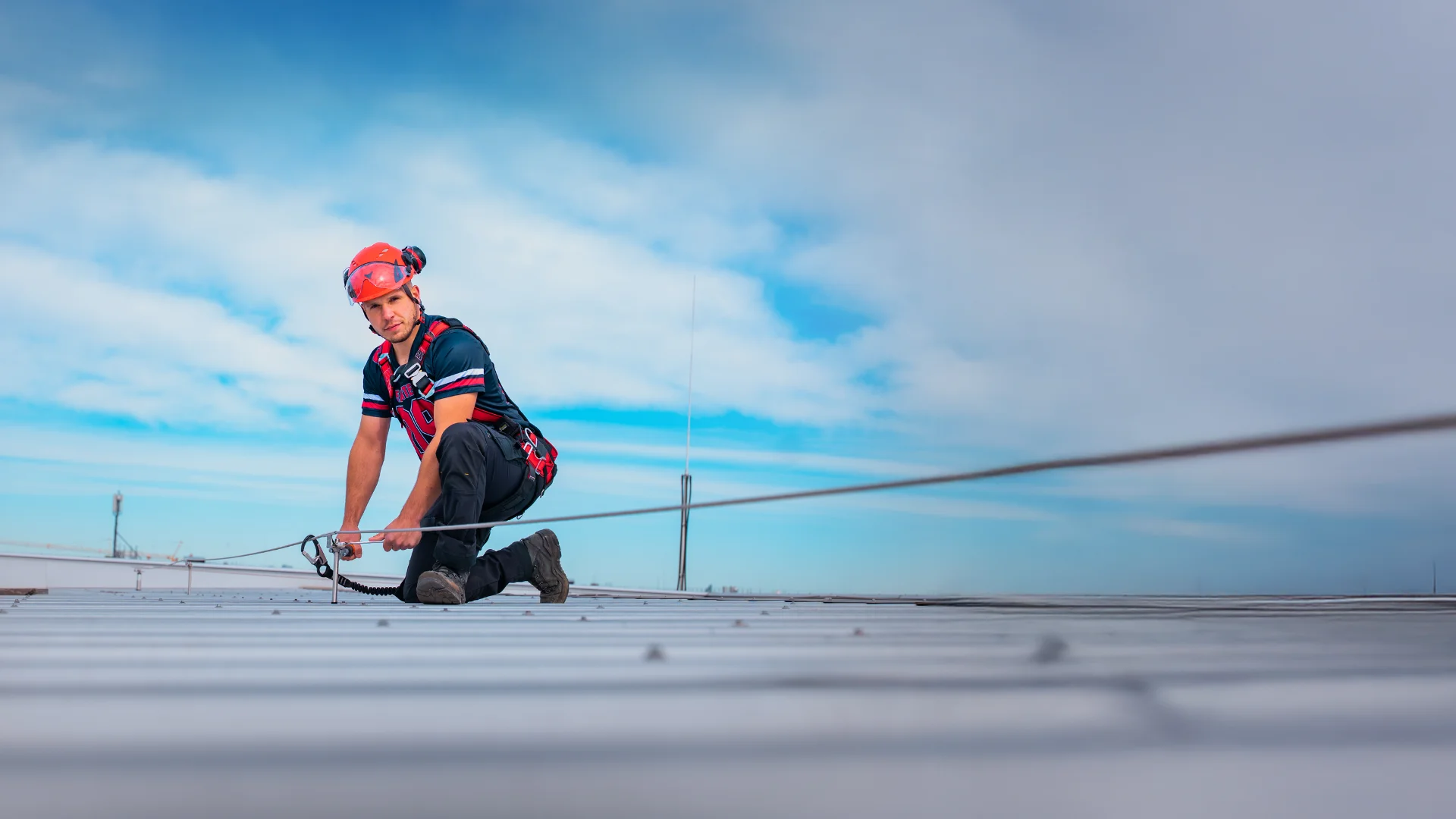 Monteur von EASiTec mit Helm und Sicherheitsgeschirr installiert Absturzsicherung auf Flachdach unter bewölktem Himmel kniend am Seilsystem.